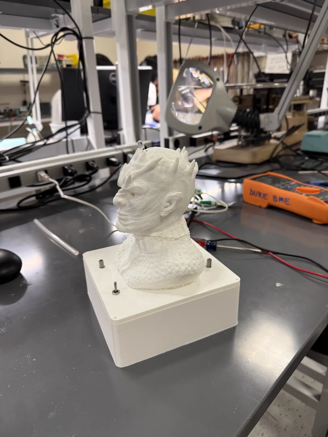 The printed enclosure in profile on a lab bench: a white 3D-printed base with visible layer lines, M3 posts standing at the corners, and the printed figure mounted on the lid.